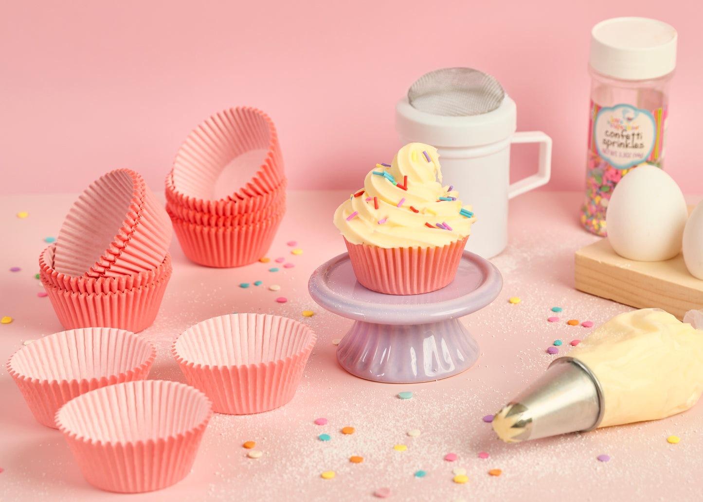 Pink cupcake cases styled with a decorated cupcake piping bag sprinkles eggs baking ingredients for cupcake decorating and home baking