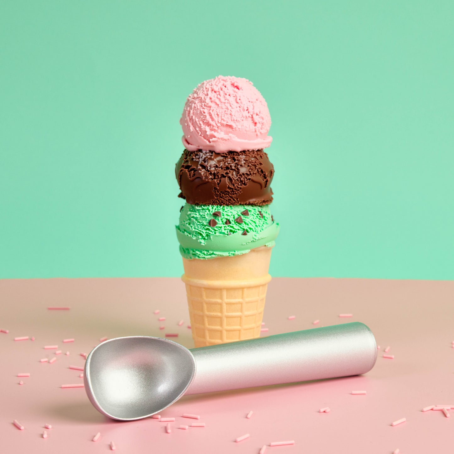 Silver ice cream scoop beside a triple scoop ice cream cone with strawberry chocolate and mint choc chip ice cream on a pastel pink surface with sprinkles and a mint background