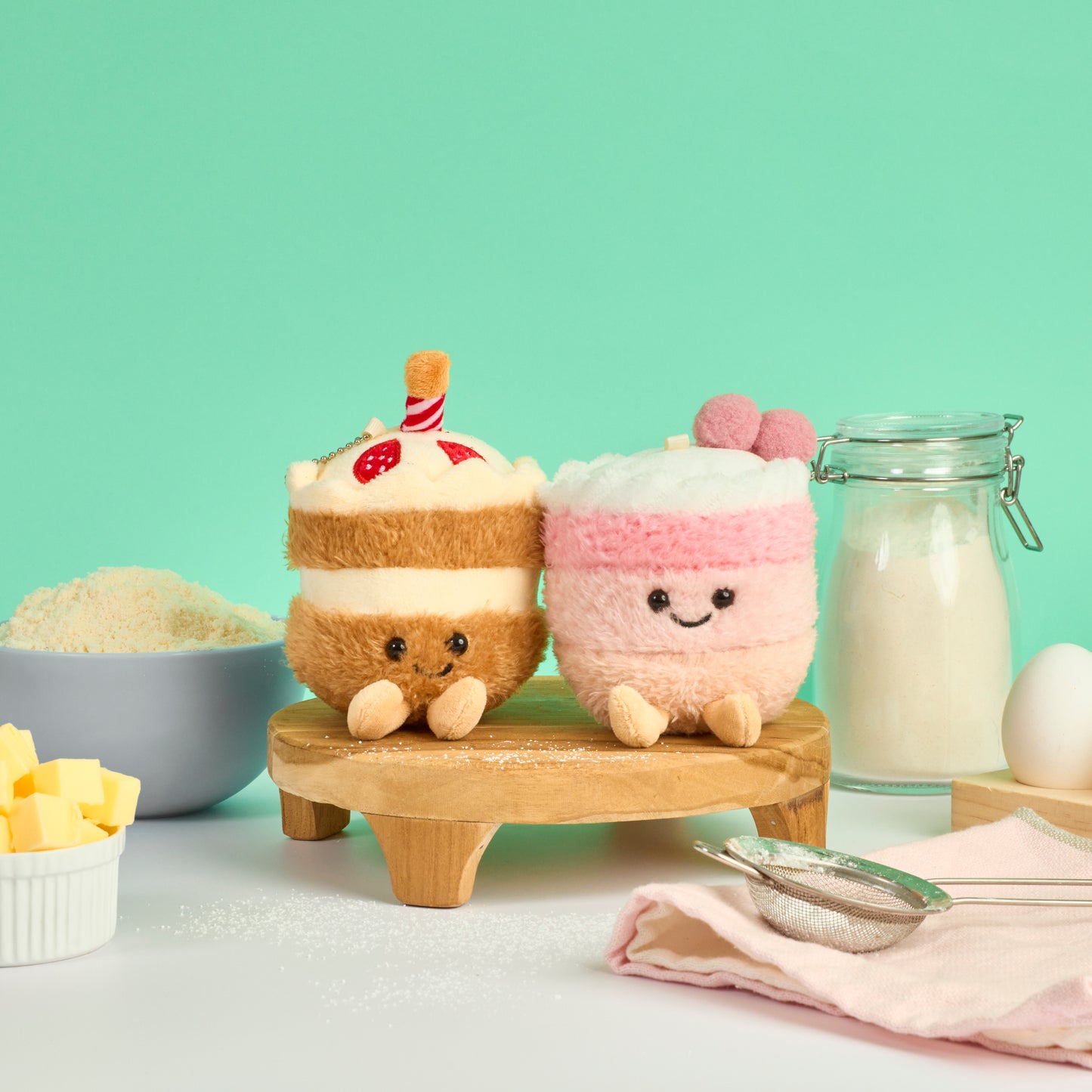Chocolate cake and pink strawberry cake plush keychains displayed with baking ingredients flour butter and eggs in a pastel kitchen setting