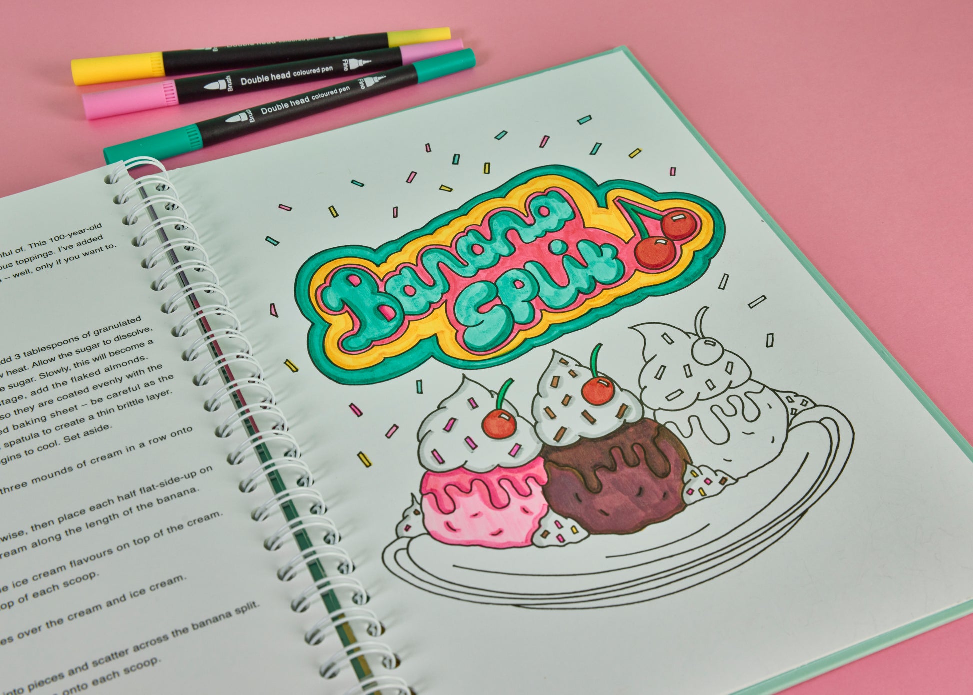Coloured banana split dessert illustration in a spiral bound colouring cookbook with dual tip brush pens on a pink background