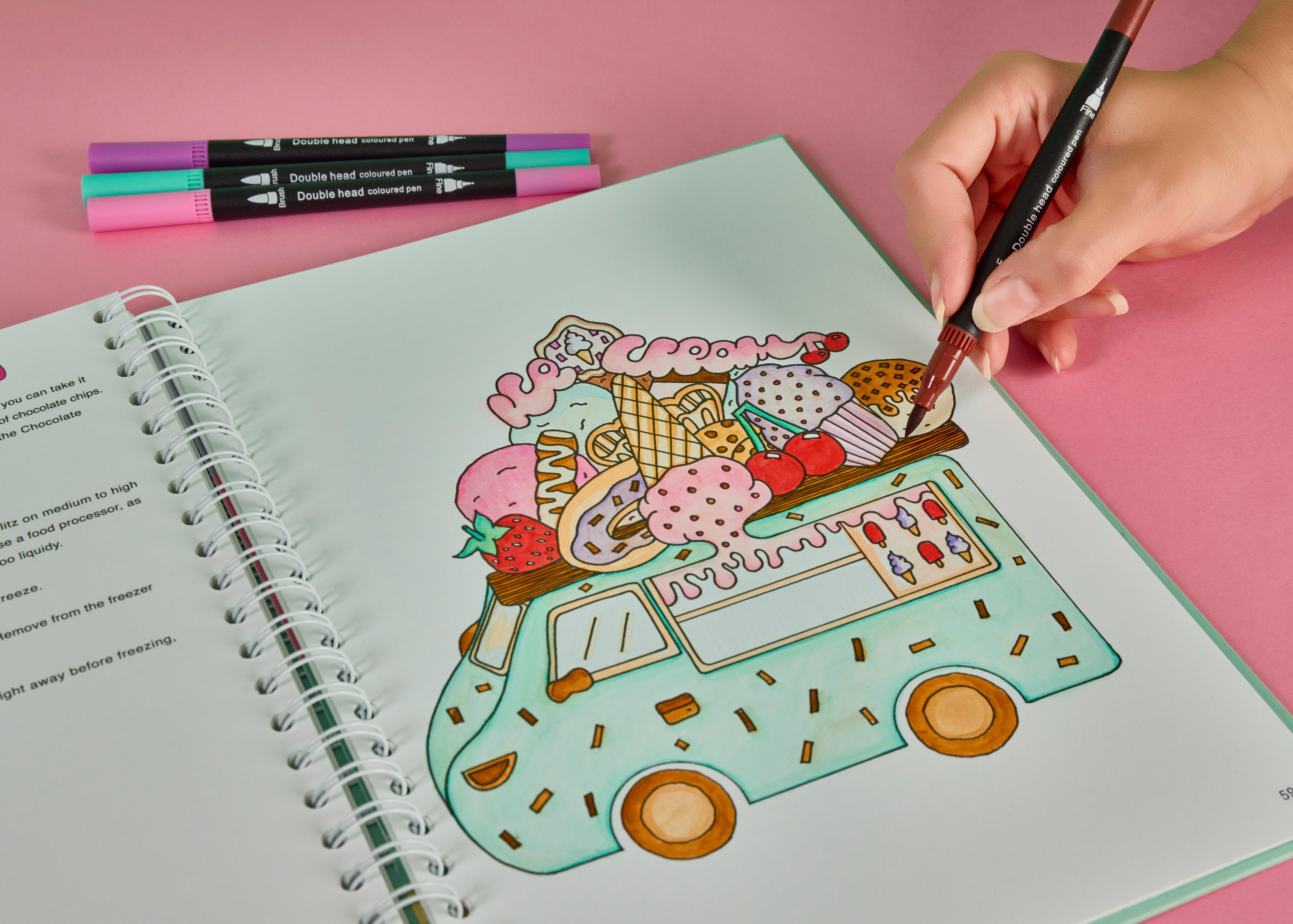 Person using dual tip brush pens to colour an ice cream van dessert illustration in a spiral bound colouring cookbook on a pink background