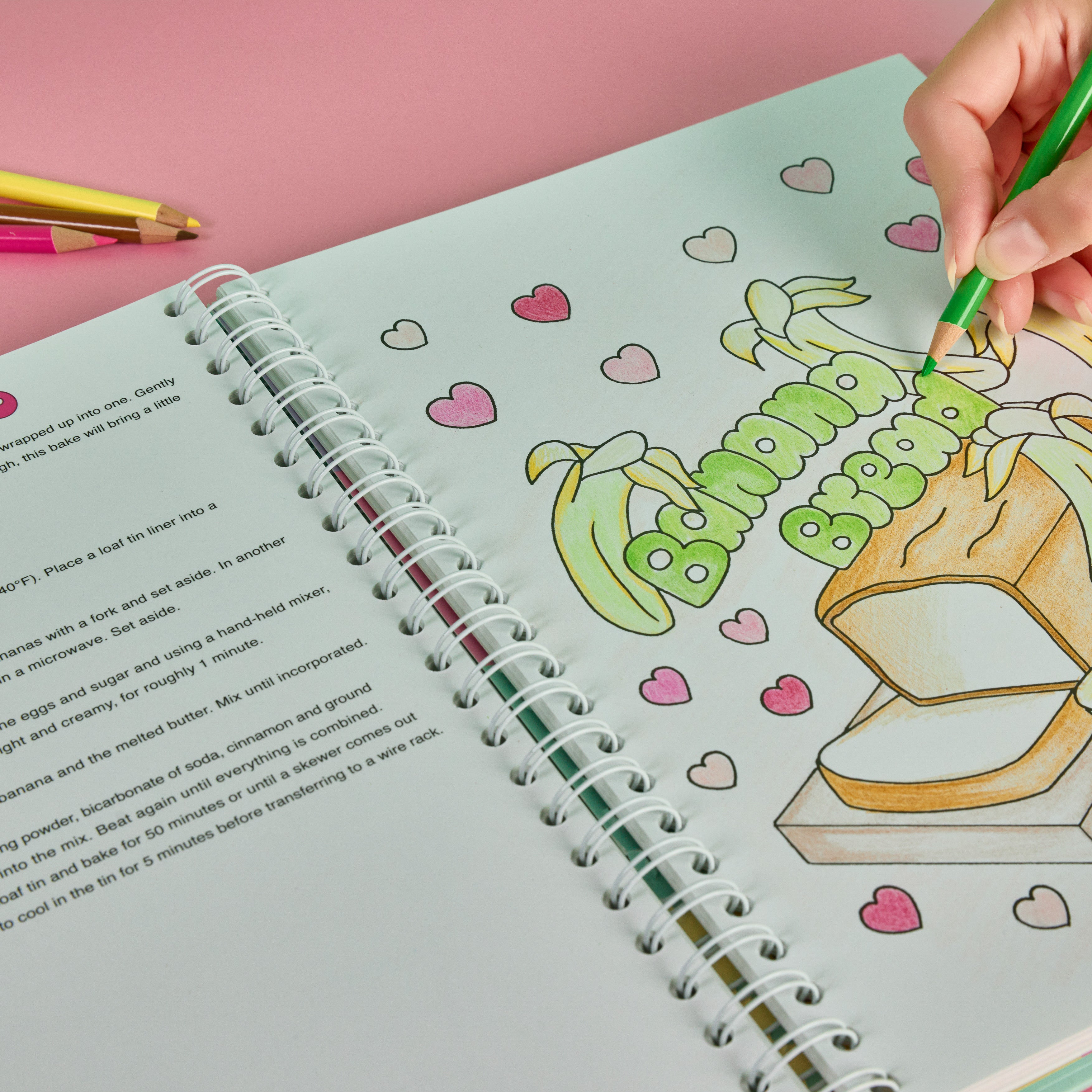 Sweet Colouring Cookbook  book with a cartoon banana cake and hearts, being coloured with a green oil pencil.