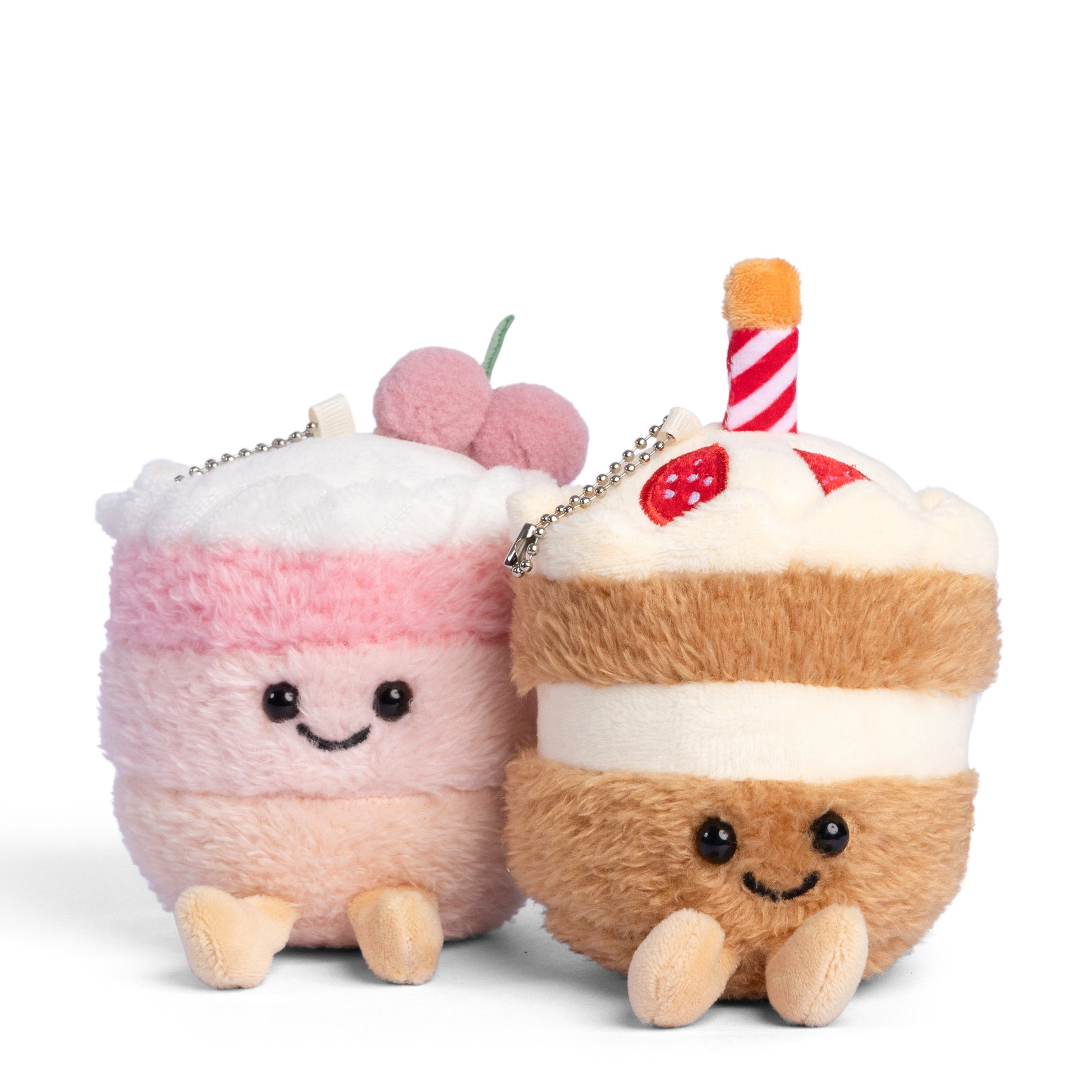 Two soft dessert plush keychains featuring a pink layered cake with cherries and a smiling strawberry cake slice with whipped cream and striped straw detail