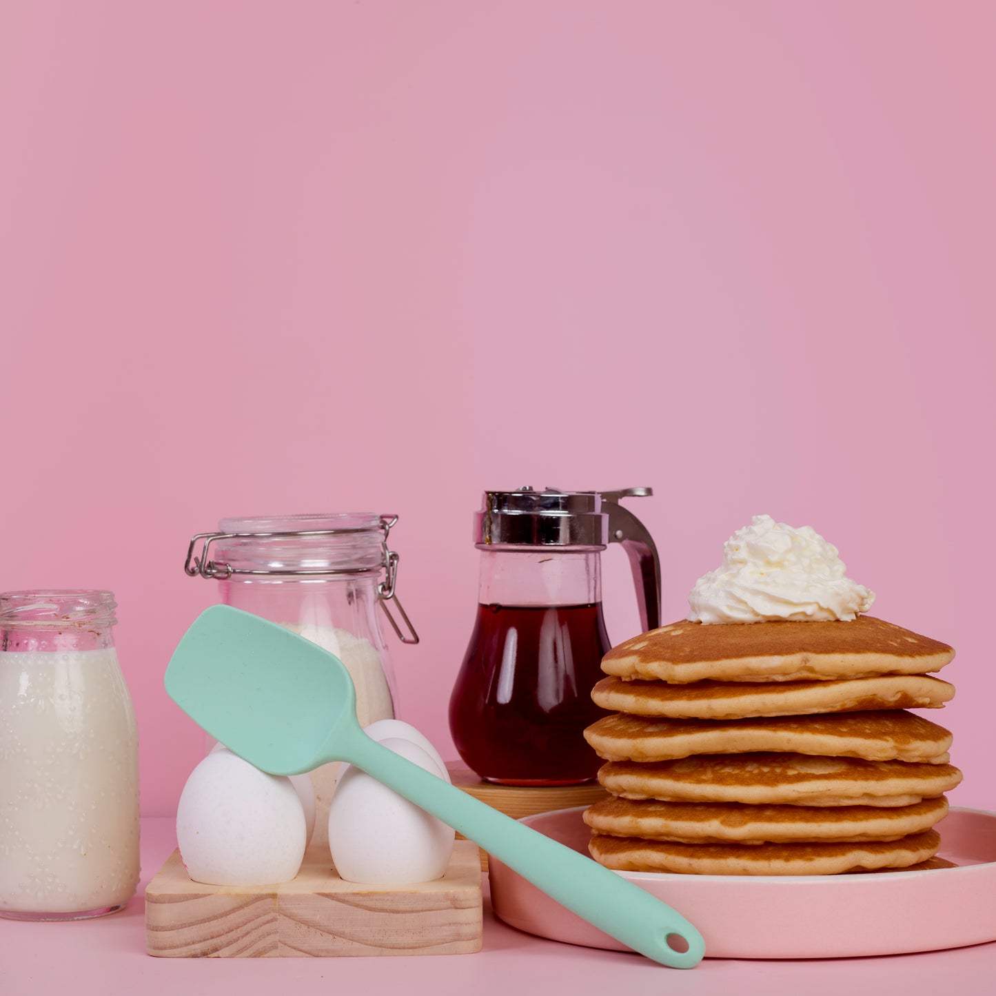 Mint green silicone spatula for mixing scraping folding batter cake decoration and home baking lifestyle shot with stacked pancakes maple syrup in a jar eggs and flour jars on a pink backfround