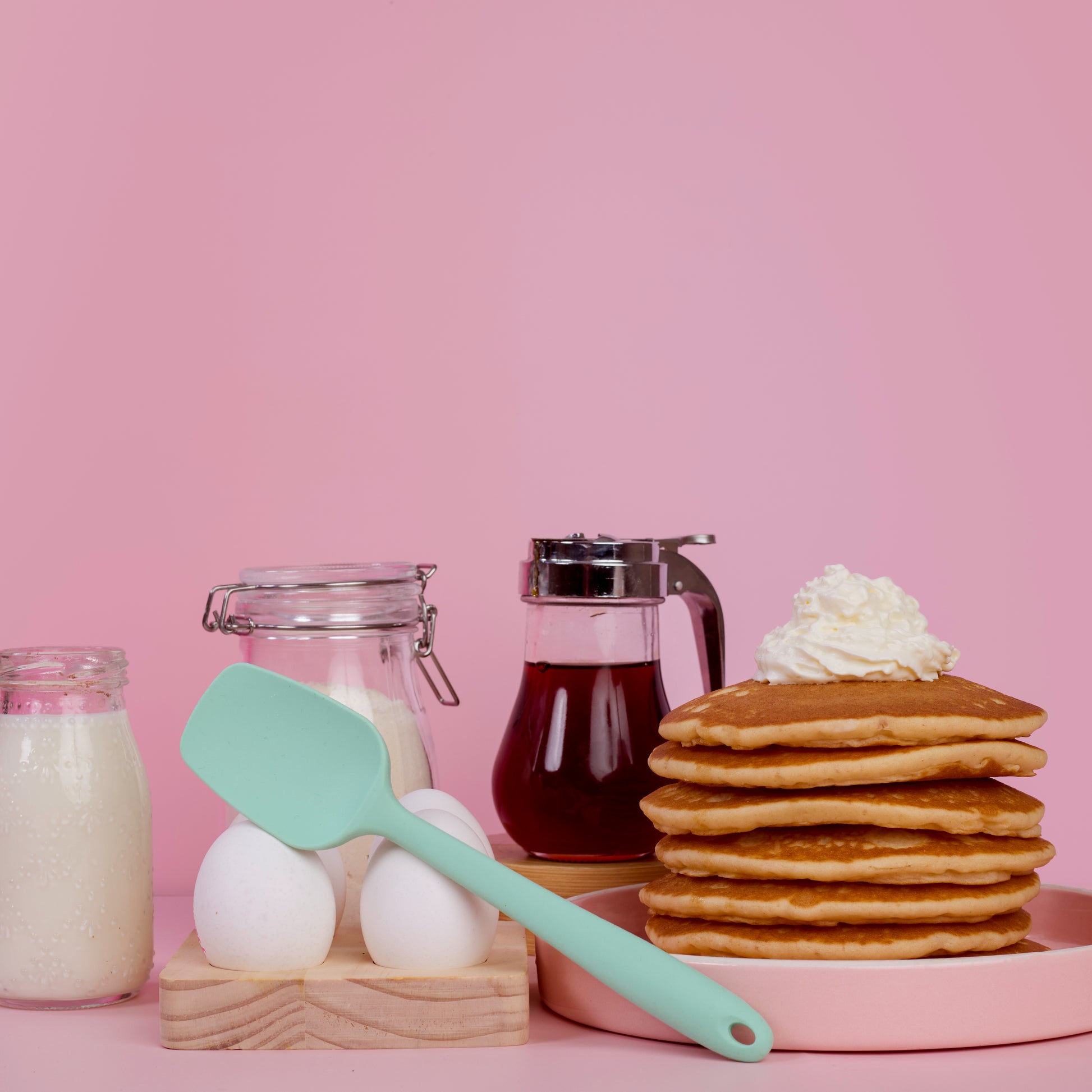 Mint green silicone spatula for mixing scraping folding batter cake decoration and home baking lifestyle shot with stacked pancakes maple syrup in a jar eggs and flour jars on a pink backfround