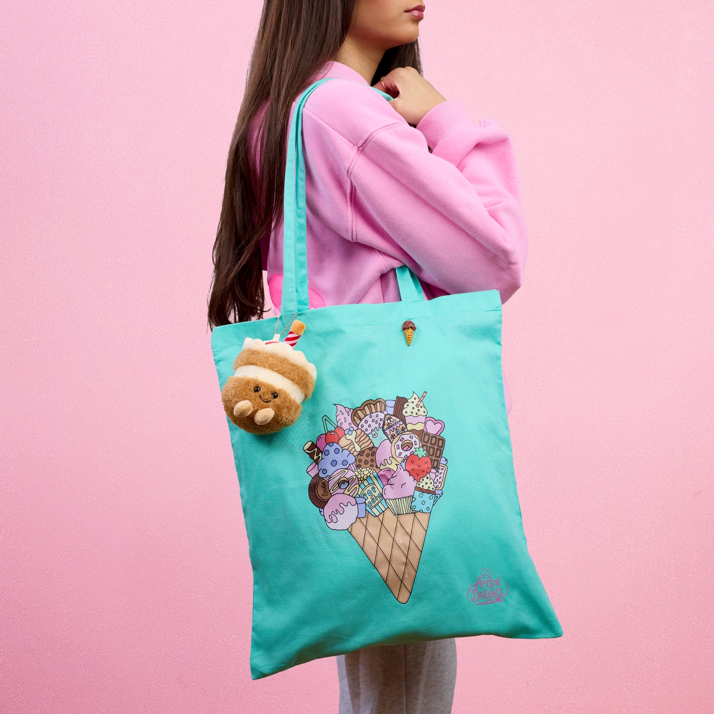 Mint green tote bag with an illustrated ice cream cone filled with desserts and sweet treats with a ice cream plushy keychain and chocolate ice cream pin brooch on a pink background