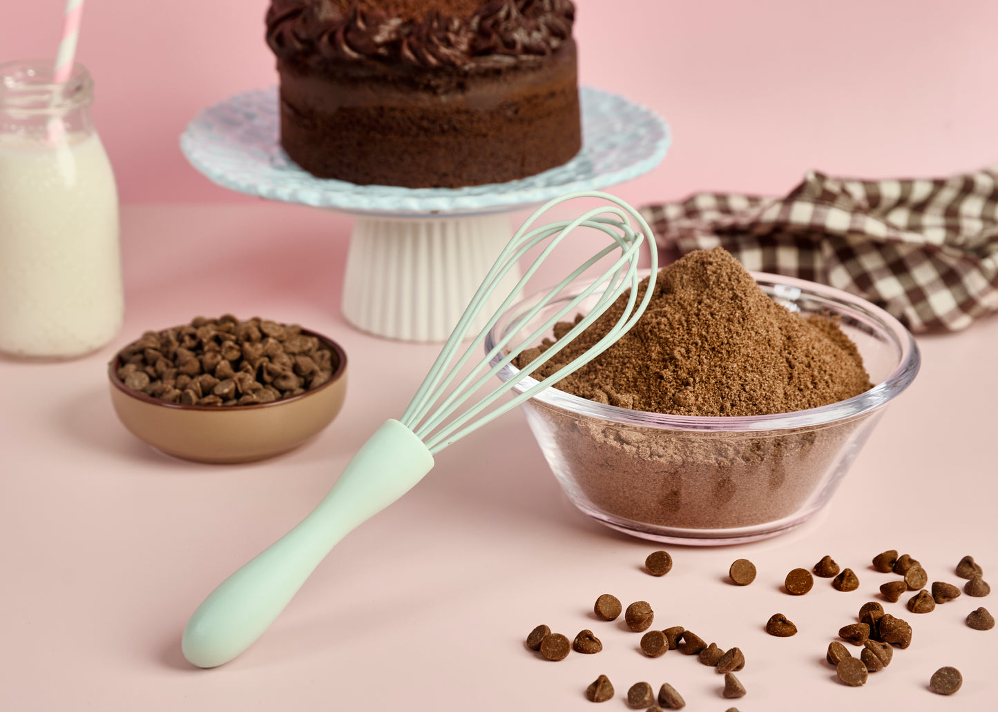 Mint green silicone whisk beside cocoa powder chocolate chips milk and chocolate cake ingredients styled for baking cake making and homemade dessert recipes