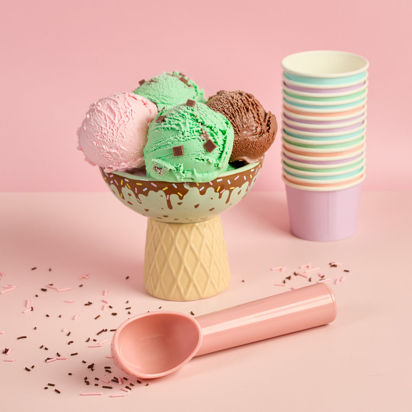 Pink ice cream scoop beside pastel ice cream bowl filled with mint chocolate chip ice cream strawberry ice cream and chocolate ice with scattered sprinkles ice cream dessert scoop gelato scoop sorbet pastel kitchen accessory ice cream serving scoop
