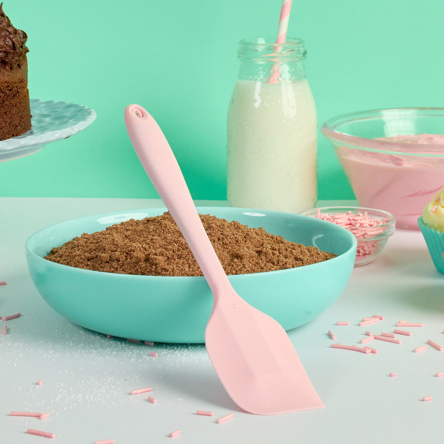 Pink silicone spatula beside a bowl of chocolate cake mixture with milk sprinkles and baking ingredients ideal fro mixing batter scraping bowls folding ingredients and homemade baking 