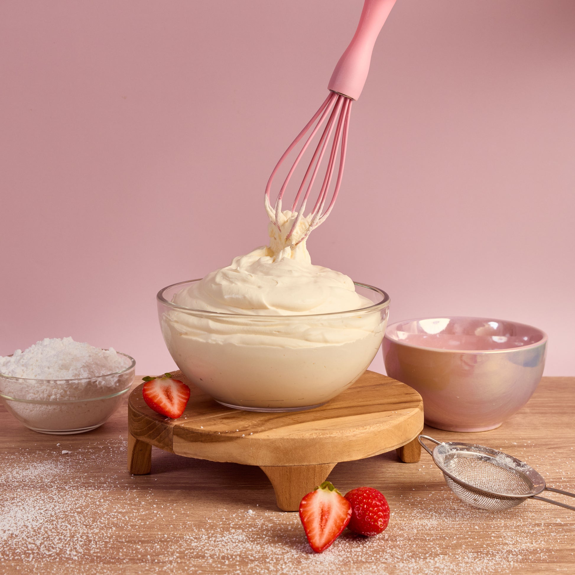 Pink Silicone whisk lifting whipped cream from a glass mixing bowl with whipped cream strawberries icing sugar and baking ingredients ideal fro whipping cream mixing batter frosting and homemade dessert recipes