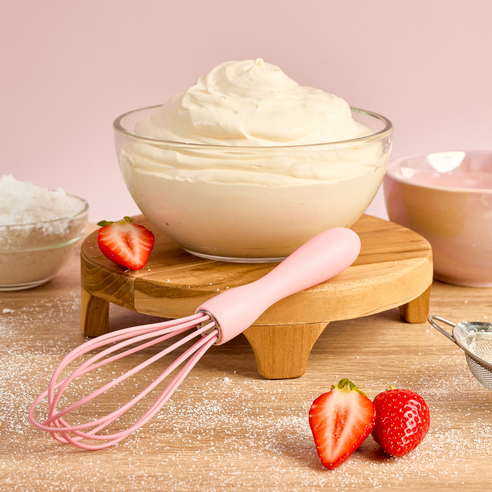 Pink Silicone whisk lifting whipped cream from a glass mixing bowl with whipped cream strawberries icing sugar and baking ingredients ideal fro whipping cream mixing batter frosting and homemade dessert recipes