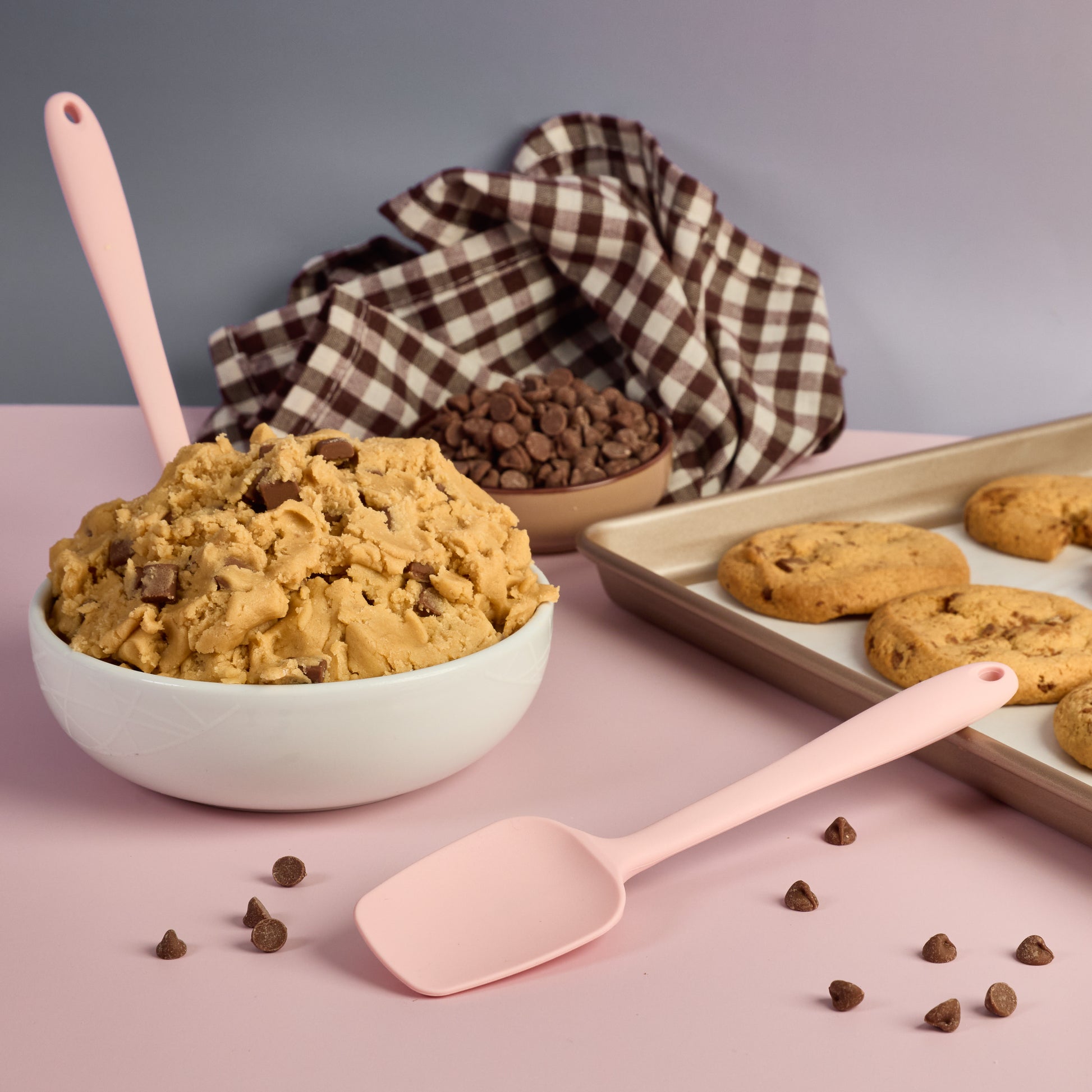 Pink Silicone spoon with chocolate chip cookie dough chocolate chips and freshly baked cookies ideal for mixing batter scraping bowls folding ingredients and homemade baking recipes chequered brown and white cloth 