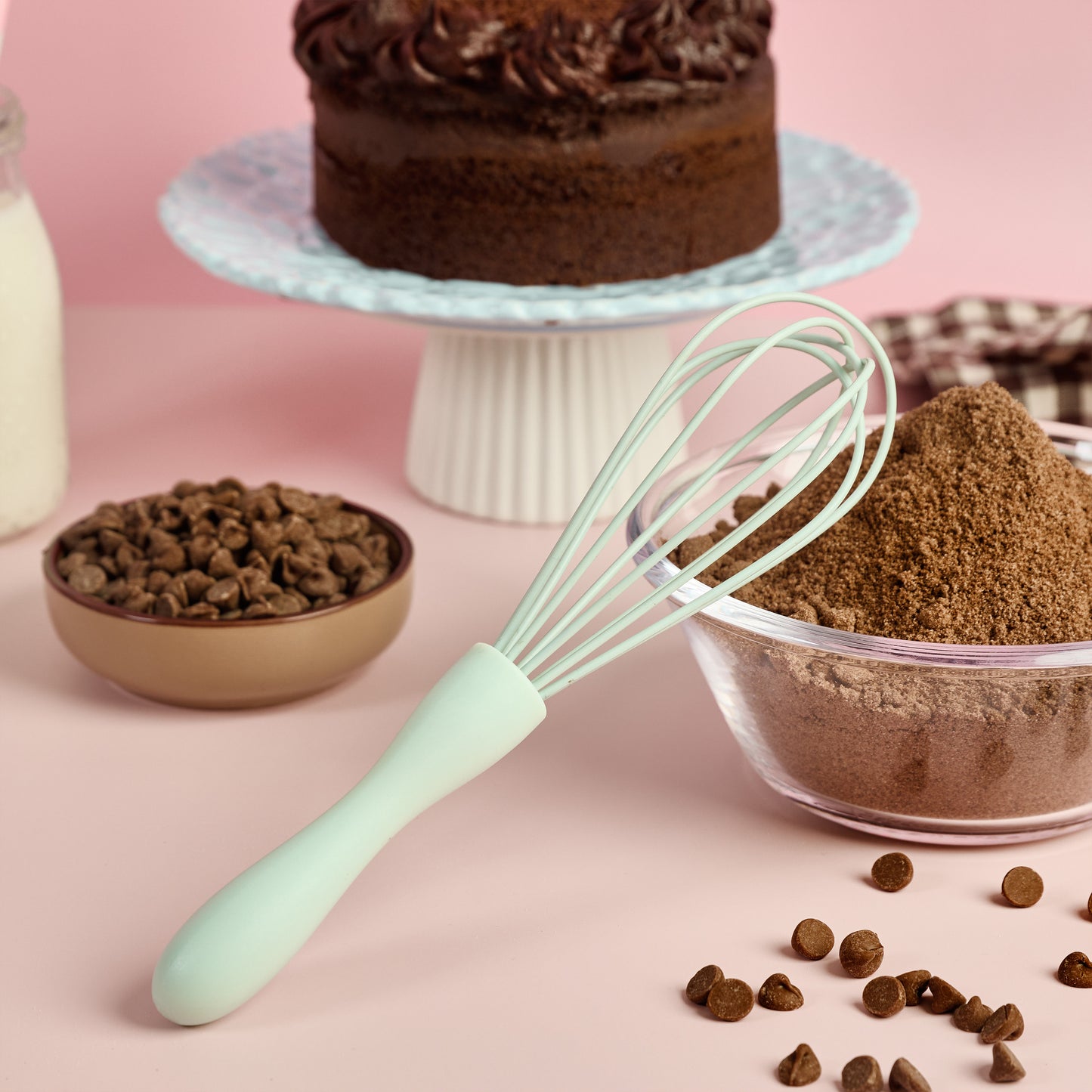 Mint green silicone whisk beside cocoa powder chocolate chips milk and chocolate cake ingredients styled for baking cake making and homemade dessert recipes