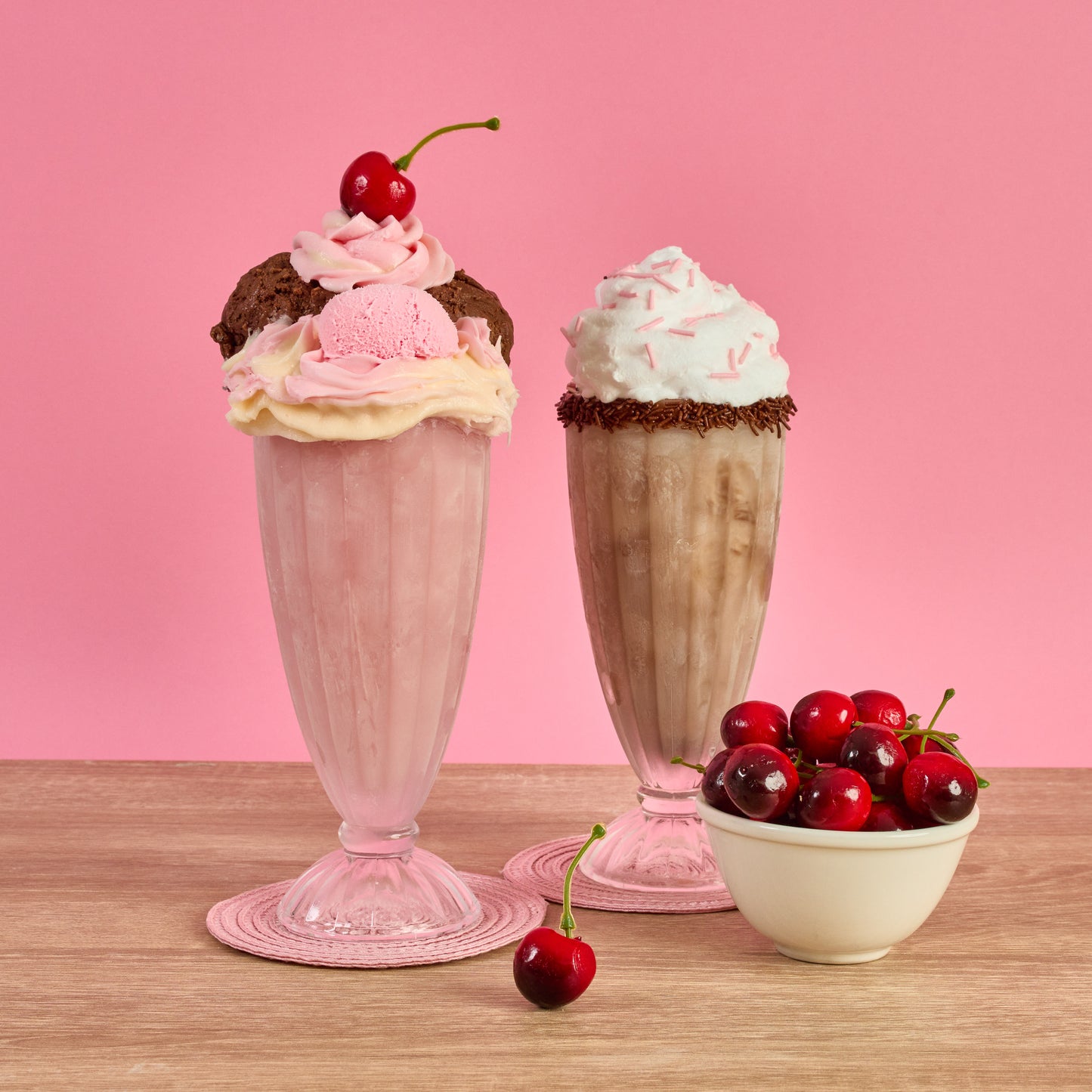 Two ice cream sundaes with pink and chocolate flavors, accompanied by a bowl of cherries on a pink background.
