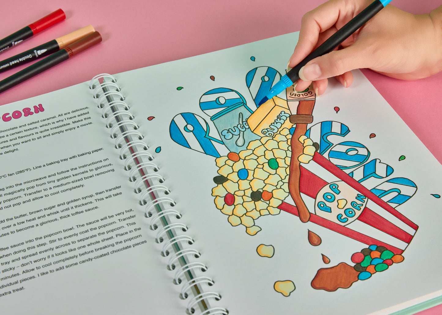 Hand colouring a toffee popcorn illustration in the Sweet Colouring Cookbook by Artful Delights using dual-tip colouring pen on a pastel pink background