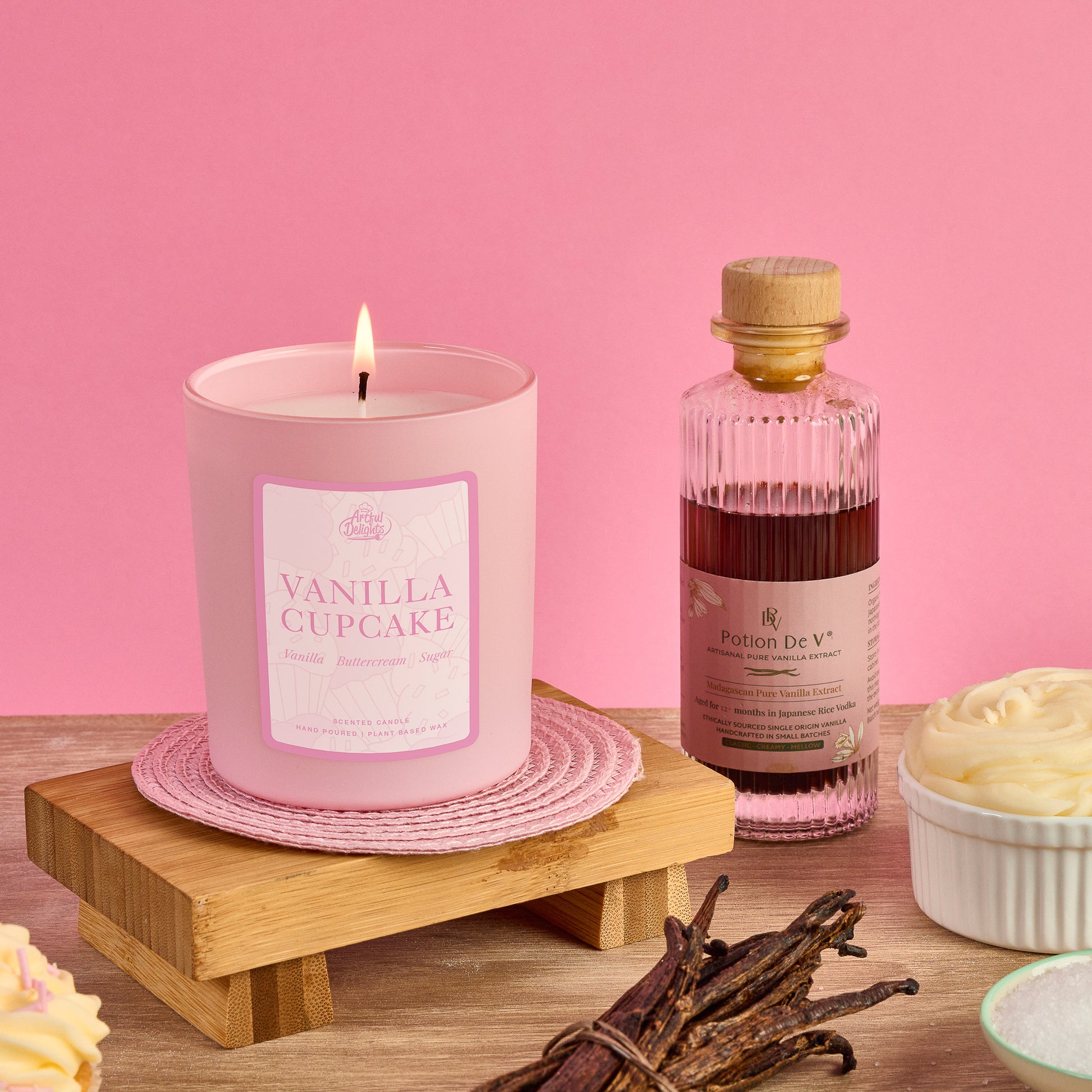 Lit vanilla cupcake scented candle in a pastel pink glass vessel style with vanilla pods, cupcake frosting and vanilla extract in a glass bottle on a wooden display against a pink background.