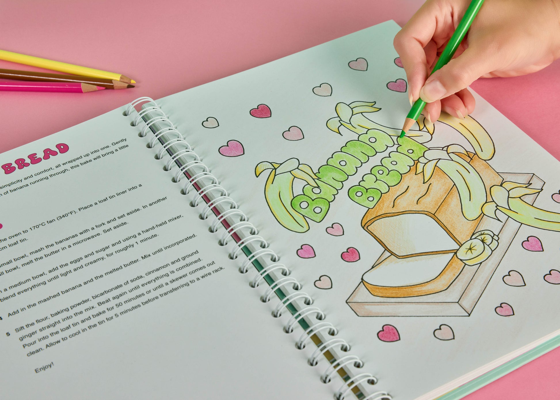 Sweet Colouring Cookbook by Artful Delights, book open to a page with a drawing of Banana bread recipe and drawing and a hand holding a green pencil with 3 pencils in background