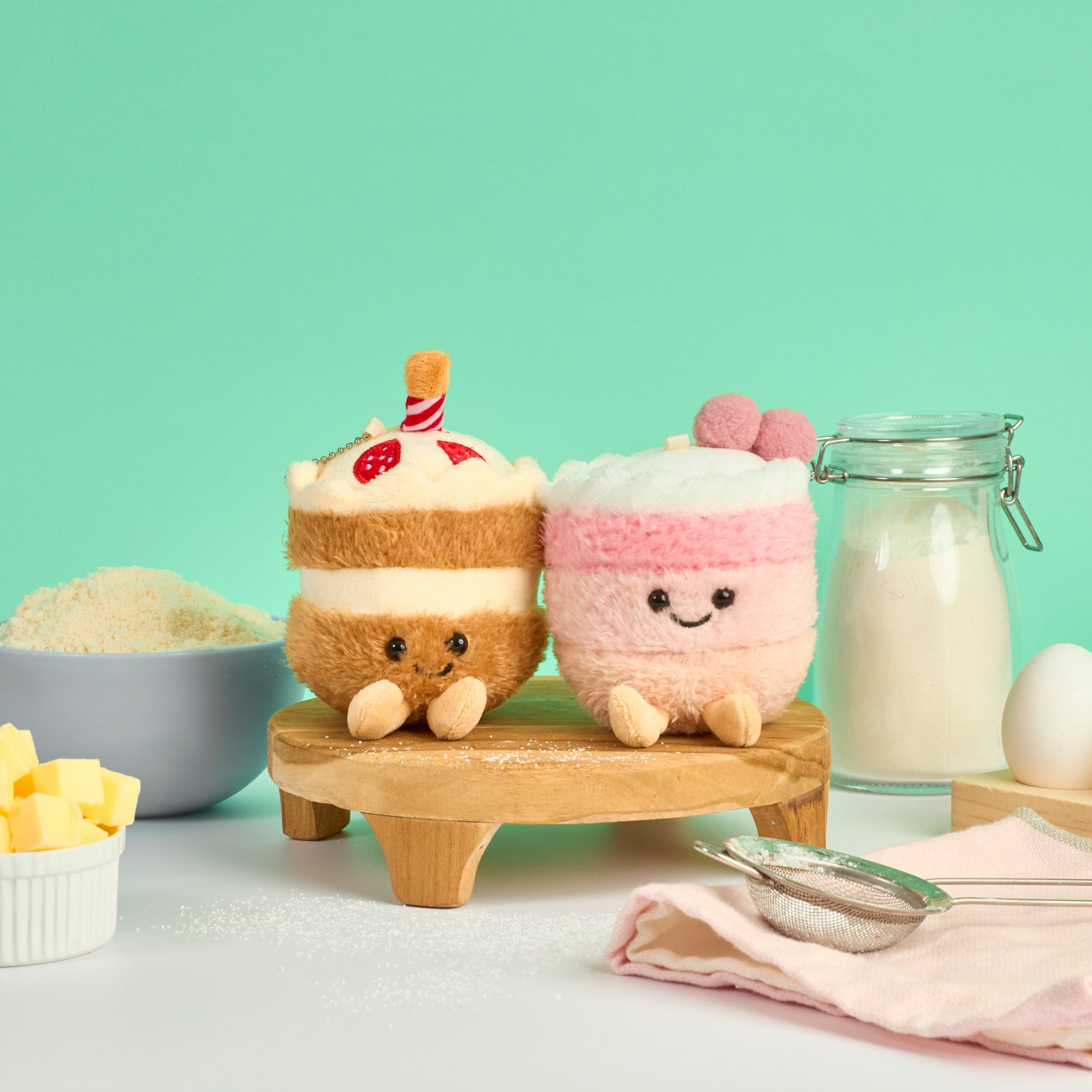 Chocolate cake and pink strawberry cake plush keychains displayed with baking ingredients flour butter and eggs in a pastel kitchen setting