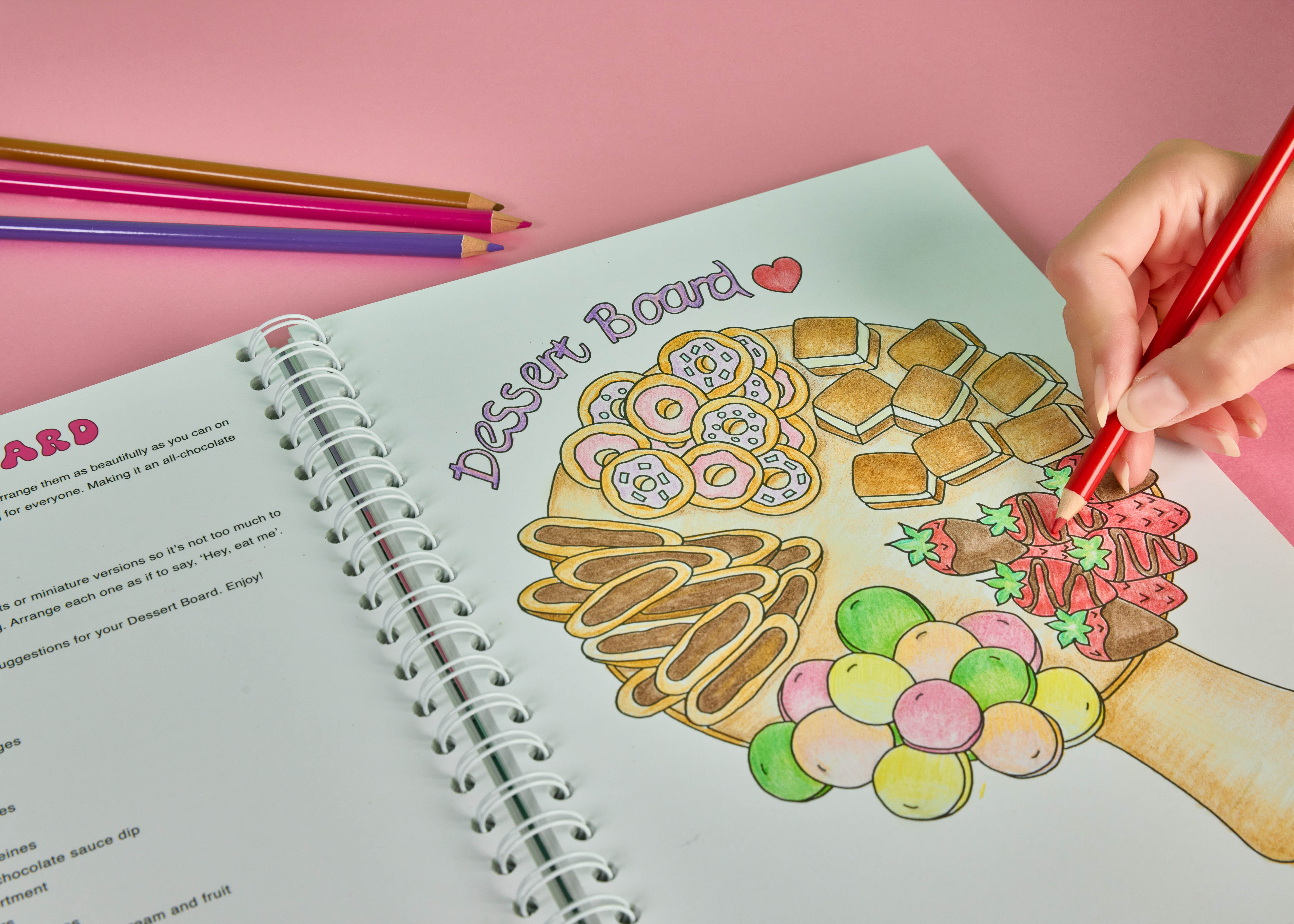 Person colouring  a dessert board illustration inside the Sweet Colouring Cookbook using coloured pencils featuring dessert themed colouring activities and creative colouring themed page being coloured by a hand holding a red pencil