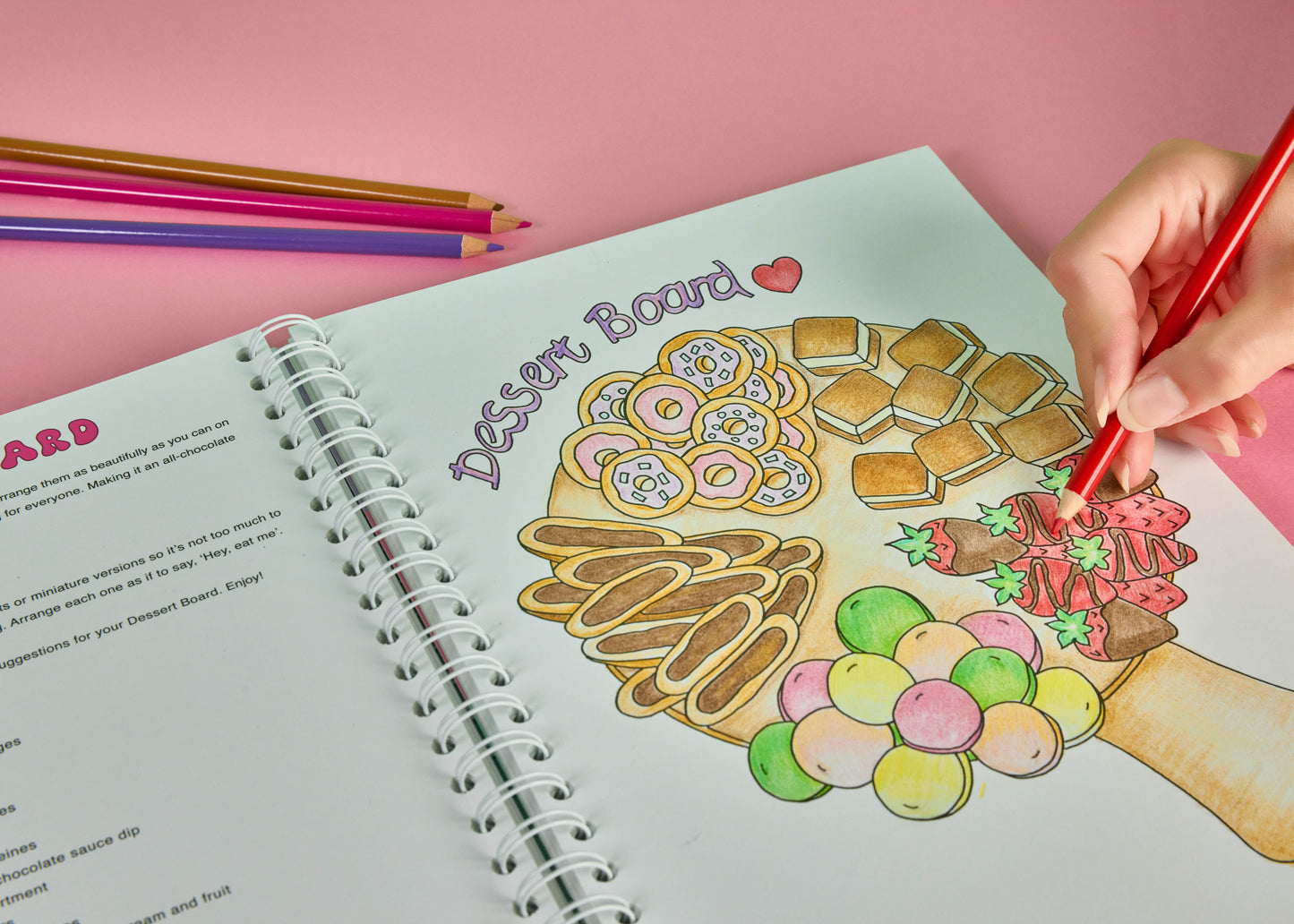 Person colouring  a dessert board illustration inside the Sweet Colouring Cookbook using coloured pencils featuring dessert themed colouring activities and creative colouring themed page being coloured by a hand holding a red pencil