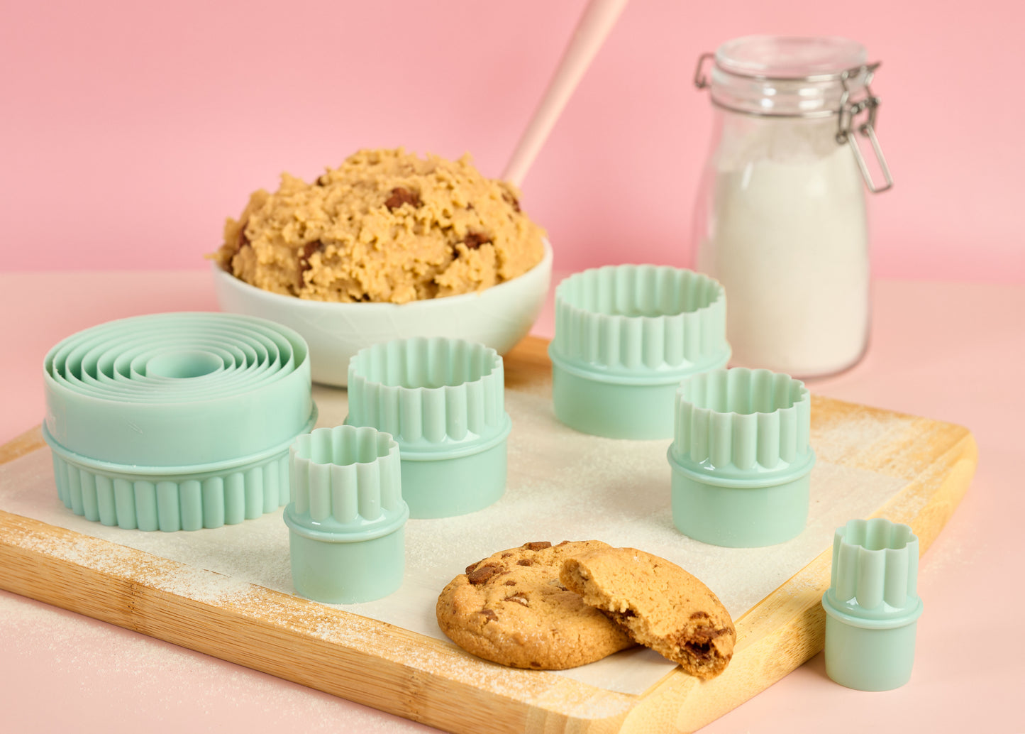 Mint green cookie cutter set with smooth and fluted edges arranged on a wooden board with cookie dough cookies and flour jar