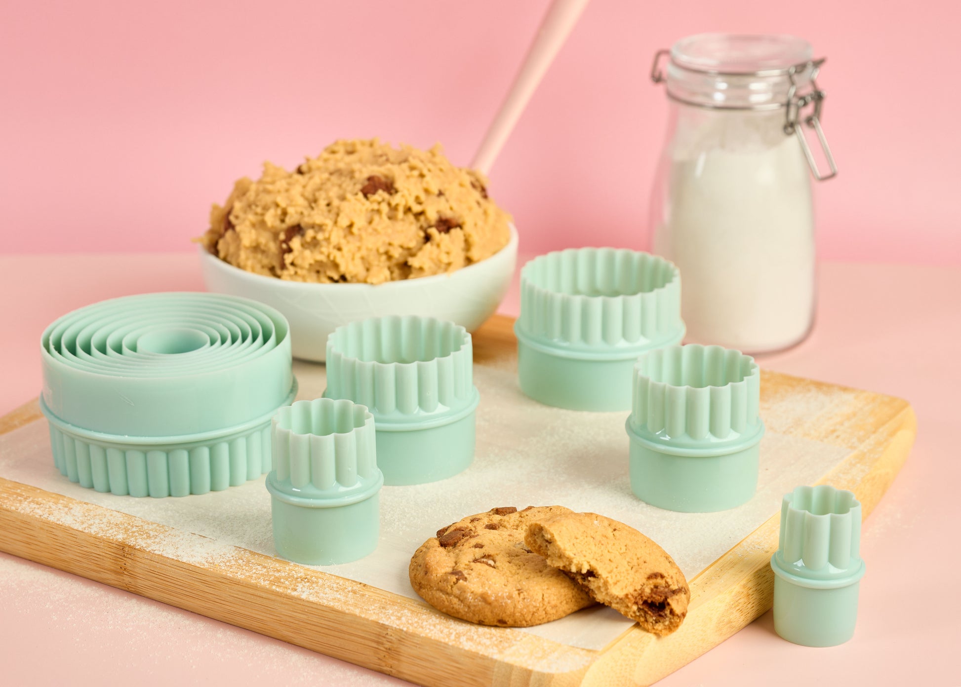 Mint green cookie cutter set with smooth and fluted edges arranged on a wooden board with cookie dough cookies and flour jar