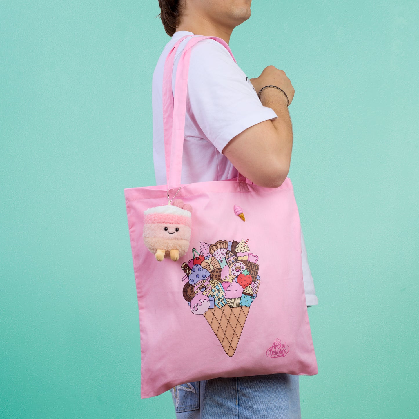 Pink tote bag with an illustrated ice cream cone filled with desserts and sweet treats with a ice cream plushy keychain and chocolate ice cream pin brooch on a mint green background