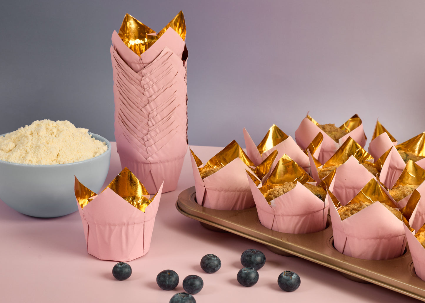 Pink and gold muffin cases filled with blueberry muffins on a baking tray with scattered blueberries and a blue bowl filled with cake mix