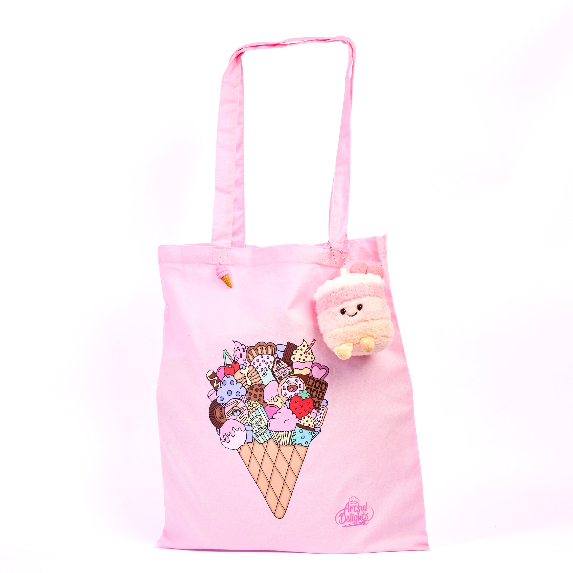 Pastel pink tote bag featuring and illustrated ice cream cone dessert design with a pink cake plushy keychain attached and pink brooch/pin on a  white background