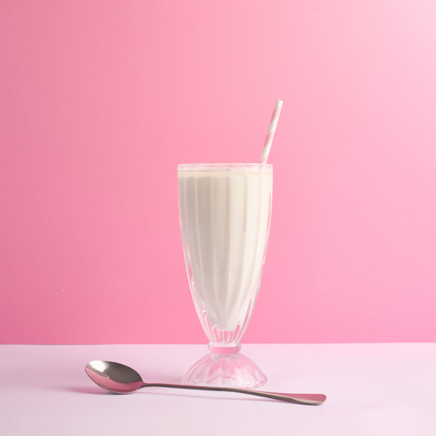 Glass of milkshake with a pink and white stripped straw on a pink background in a tall sundae glass with a long stem silver spoon.