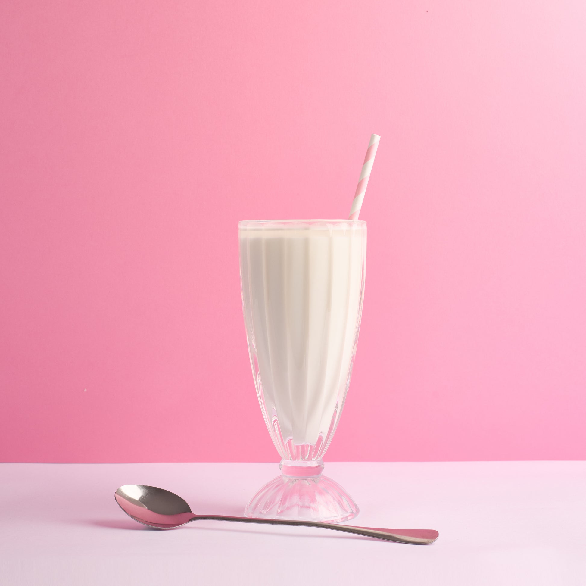 Glass of milkshake with a pink and white stripped straw on a pink background in a tall sundae glass with a long stem silver spoon.